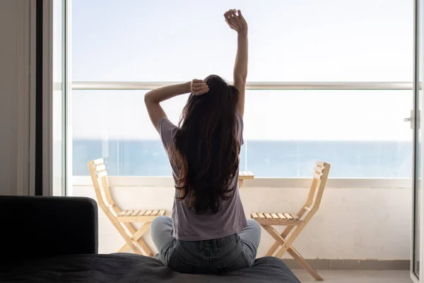 A young woman is sitting on sofa near balcony and stretching in the morning — Stock Photo, Image