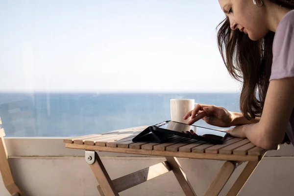 A young woman in headphones is working on tablet on the balcony near the sea — Stock Photo, Image