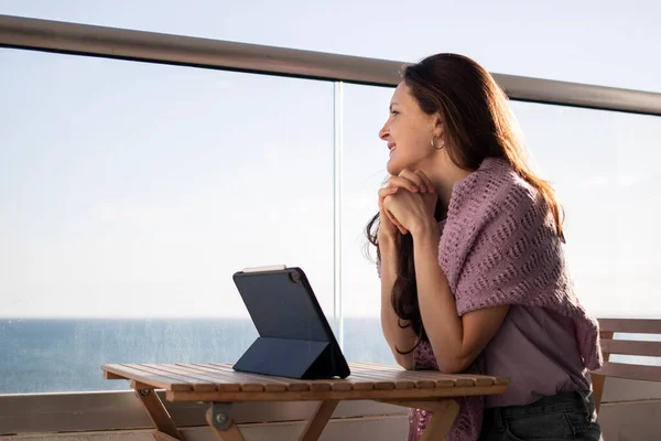 Genç bir kadın tabletle balkonda oturuyor ve deniz kıyısına bakıyor. — Stok fotoğraf
