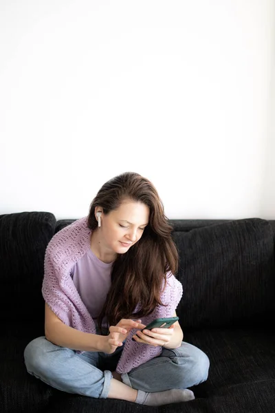 A young woman in headphones is sitting on a sofa and using her phone — Stock Photo, Image