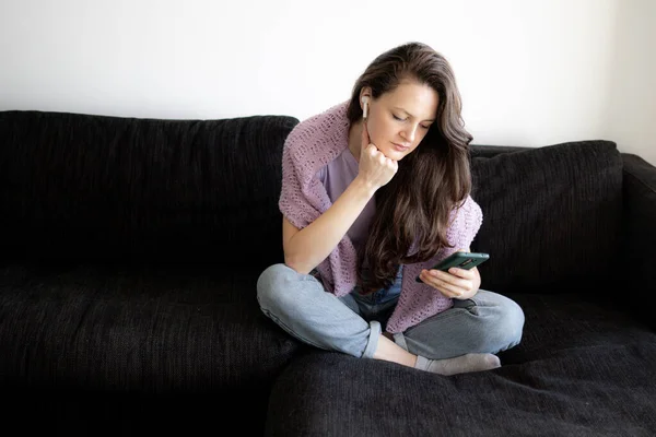 A woman in headphones is sitting on a couch and bored using her phone — Stock Photo, Image