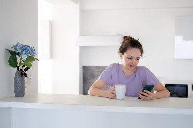Genç bir kadının mutfakta telefonunu kullanıp çayını içmesinin sabah rutini.