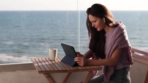 Ung Kvinna Hörlurar Arbetar Tablett Balkongen Nära Havet Med Kopp — Stockvideo