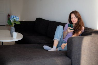 Genç bir kadın evdeki kanepede oturuyor ve telefonunu kullanıyor.. 