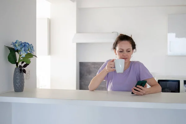 The morning routine of a young woman using her phone in the kitchen — Stock Photo, Image