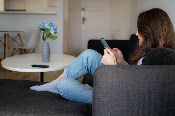 En ung kvinna sitter i en soffa och använder sin mobil. — Stockfoto