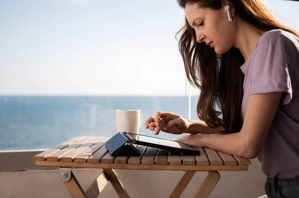 Kulaklıklı genç bir kadın deniz kenarındaki balkondaki tablette yazı yazıyor. — Stok fotoğraf