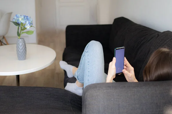 A young woman is lying on a sofa and using her phone — Stock Photo, Image