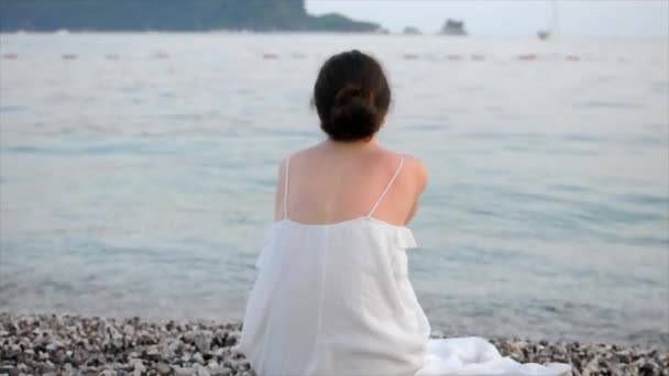 Een jonge vrouw zit aan het strand en gooit een kiezelsteen in de zee. — Stockvideo