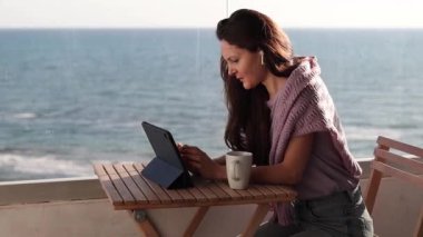 Dijital tablet üzerinde çalışan, terasta stili olan çay içip denize bakan bir kadının yan görüntüsü.