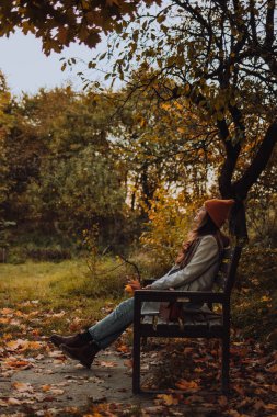 Genç bir kadın tek başına bankta oturuyor ve sonbahar sezonunda yukarı bakıyor.