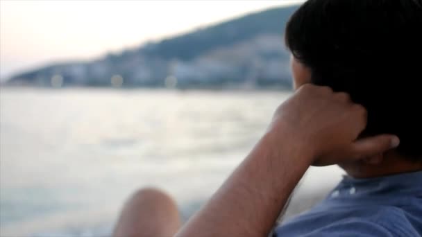Jeune homme ennuyé est assis à la plage et regardant la mer — Video
