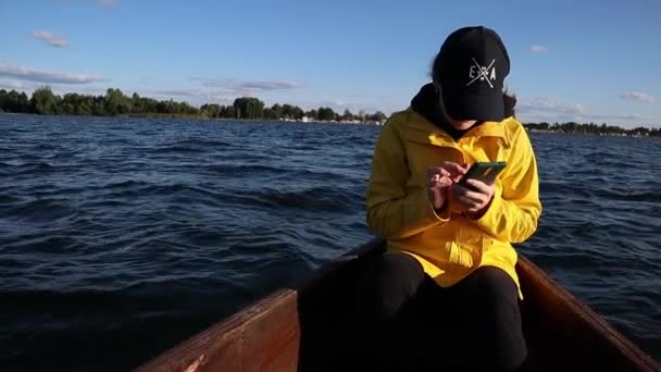 Mujer joven con impermeable amarillo y gorra sentada en un barco y mirando el teléfono — Vídeo de stock