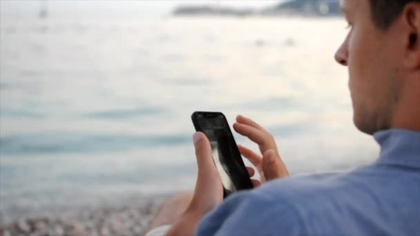 Νεαρός άνδρας χρησιμοποιεί το smartphone του ενώ κάθεται στην παραλία κοντά στη θάλασσα — Αρχείο Βίντεο