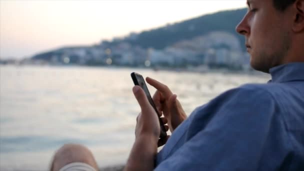 Jeune homme utilise son smartphone tout en étant assis à la plage près de la mer — Video