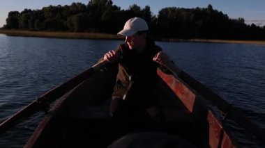 Genç adam, güneşli bir günde, tahta küreklerle bir gölde kürek çekiyor.