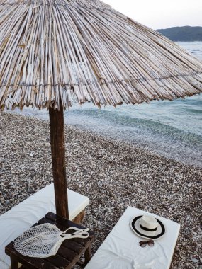 Kumsal manzaralı deniz manzaralı güneşli yataklar ve çakıl taşlı plajda bir hasır şemsiye. Yaz aksesuarları: Hasır şapka, çevre dostu ağ alışveriş çantası ve güneş gözlüğü..