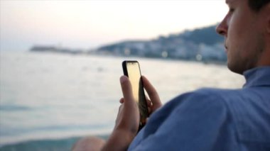 Genç adam deniz kenarında sahilde otururken akıllı telefonunu kullanıyor. Adam deniz manzarasının fotoğrafını çekiyor.