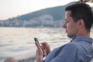 Genç bir adam deniz kenarında sahilde otururken cep telefonunu kullanıyor. Tatilde e- postaları kontrol eden kişi