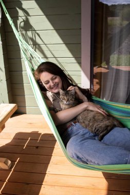 Kadın köydeki evin yakınındaki bir hamakta yatarken kedinin kafasını okşar.