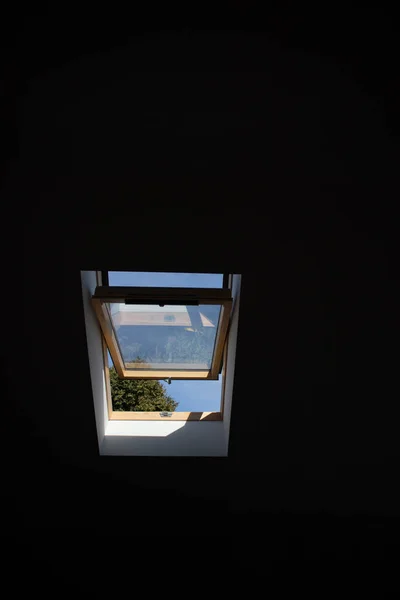 Open roof window skylight with a black background. View of the sky and trees from the window. Sunlight through the attic window. Black contrast light in the room