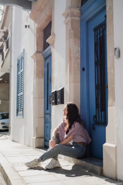 Kıbrıs Rum Kesimi 'nin Larnaka kentinde mavi kapıları ve pencereleri olan güzel bir binanın yanında oturan genç bir kadın görülüyor..