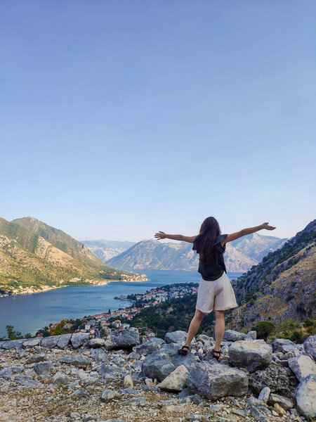 A happy young woman with open hands is standing and looking at Kotor bay with the sea and mountains view from above in Kotor, Montenegro