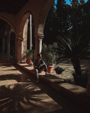 Kıbrıs Rum Kesimi 'nin Panagia Chrisopolitissa kentinde kemer ve saksı bitkilerine sahip güzel binanın yanında oturan genç bir adam görülüyor. Palmiye ağacı silueti.