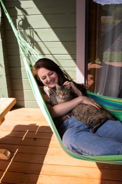 Kadın köydeki evin yakınındaki bir hamakta yatarken kedinin kafasını okşar.
