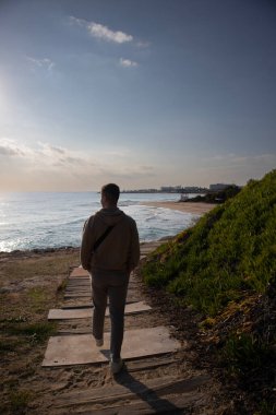 Güneşli bir günde, Belind 'li genç bir adam Kıbrıs Rum Kesimi' nin Ayia Napa kentinde, deniz kenarındaki kumlu plaja giden ahşap patikada yürüyor.