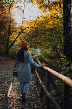 Genç bir kız sonbahar mevsiminde ahşap parkta tek başına yürüyor. Dışarıda turuncu şapkalı ve gri yün ceketli bir kadın var..