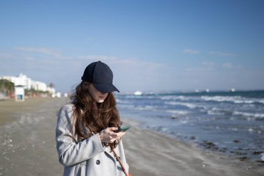 Siyah şapkalı genç bir kadın deniz kenarında akıllı telefon kullanıyor.. 