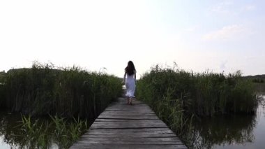 Arkadan çıplak ayaklı, beyaz elbiseli genç bir kadın göle giden tahta bir köprüde koşuyor..