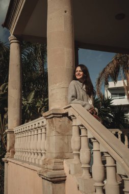 Kıbrıs Rum Kesimi 'nin Larnaka kentinde sütunları ve tropikal bitkileri olan eski binanın yanında duran ve eğlenen genç bir kadın görülüyor..