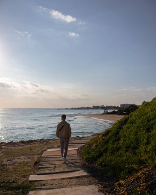 Güneşli bir günde, Belind 'li genç bir adam Kıbrıs Rum Kesimi' nin Ayia Napa kentinde, deniz kenarındaki kumlu plaja giden ahşap patikada yürüyor.
