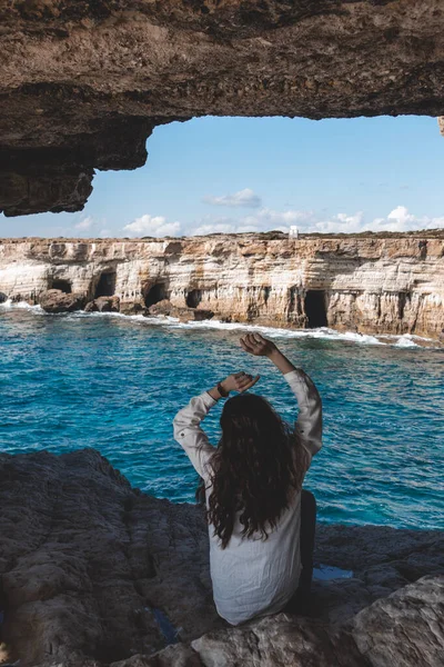 Young woman from behind is sitting in cave hole and looking at sea caves near cape Greco in national park with turquoise water sea in Ayia Napa, Cyprus.