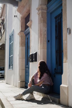 Kıbrıs Rum Kesimi 'nin Larnaka kentinde mavi kapıları ve pencereleri olan güzel bir binanın yanında oturan genç bir kadın görülüyor..