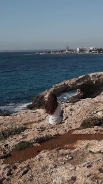 Arkadan kaygısız genç bir kadın, Kıbrıs Rum Kesimi 'nin Ayia Napa kentindeki deniz mağaralarının kenarında yalnız başına aşk köprüsünde oturuyor.