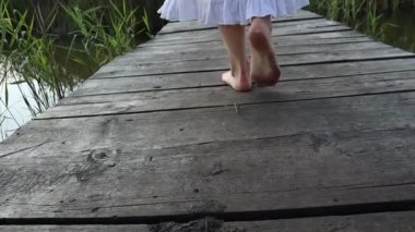 Arkadan çıplak ayaklı, beyaz elbiseli genç bir kadının yakın çekimi, göle giden ahşap bir patika köprüsünde yürüyor.