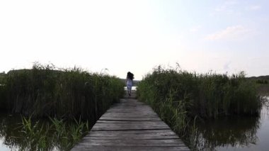 A young barefoot woman from behind in a white dress is running away on a wooden trail bridge to the lake, turning around at the edge and running back.