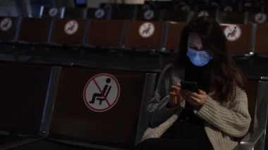 Side view of a young woman in medical mask waiting in the airport terminal and looking in her phone