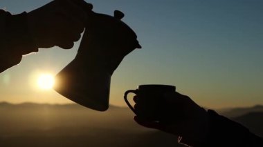 The person pouring out coffee from Moka pot into a cup outdoors in front of a sunrise. Foggy mountains background