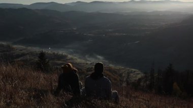 A young couple from behind is sitting in front of the beautiful mountains view and enjoying the sunrise. The woman and the man are having a break outside together.