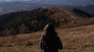 A young brunette woman from behind is standing in front of the mountains background. The girl is enjoying a beautiful sunny view of a road.