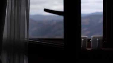 Two cups of tea on the windowsill with an opened window in front of the beautiful mountains background in foggy day