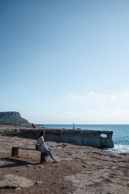 A young woman is sitting on a bench and looking at sea caves near cape Greco in a national park in Ayia Napa, Cyprus. 