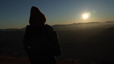 The silhouette of a young woman in a hat drinking tea from a cup outdoors in front of mountains. Cloudy hill background in the autumn season