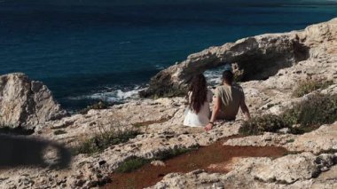 Arkadan genç bir çift kıyıda oturuyor ve Kıbrıs Rum Kesimi 'nin Ayia Napa kentindeki deniz mağarası kemer köprüsüne bakıyor.