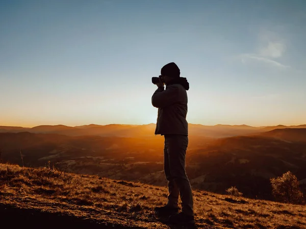 A silhouette of a man photographer taking pictures in front of autumn mountains landscape during sunrise