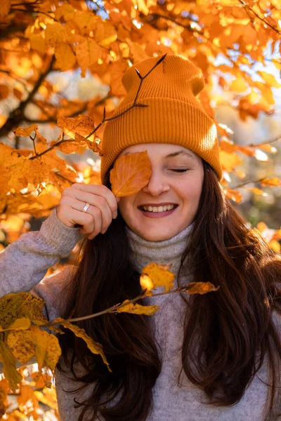 A portrait of a happy smiling woman in an orange beanie in the orange leaves. Girl with closed eyes holding a leaf in front of her face